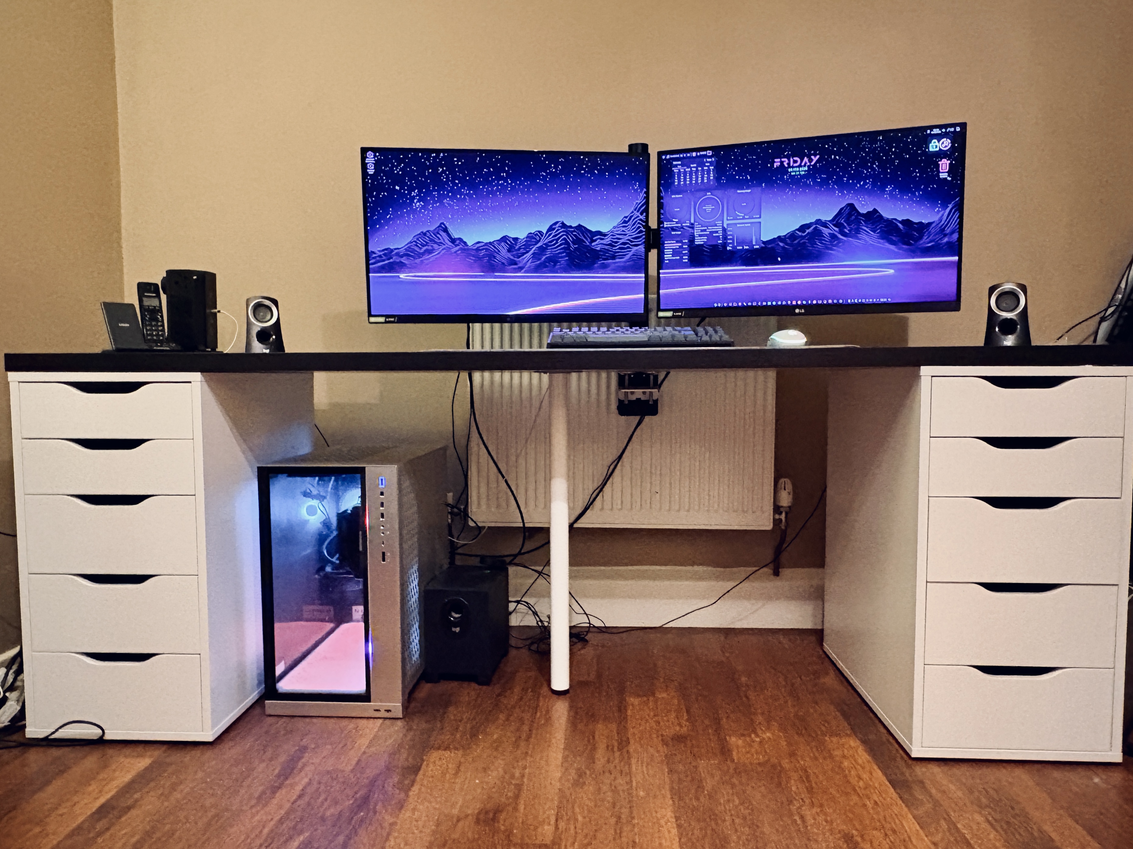 A photo depicting a desk of 2 metres in width. There are white drawers at either end, supporting the desk surface which is a darker, almost black, material. On the desk is a mechanical keyboard, two 27" monitors, a white Logitech mouse, speakers and assorted electrical gadgetry. Underneath the desk sits a large PC, encased partially in glass with RGB lighting.